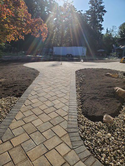 Brick pathway curves through a park with sunlight streaming. Newly planted dark soil borders the path.