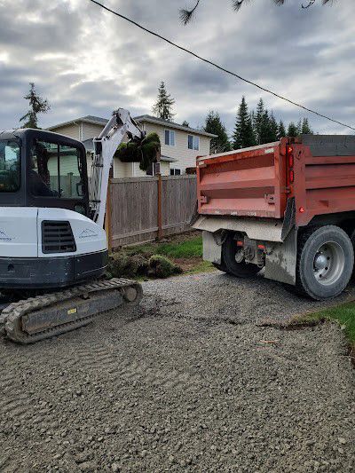 Small excavator loading dirt into an orange dump truck on a gravel driveway.