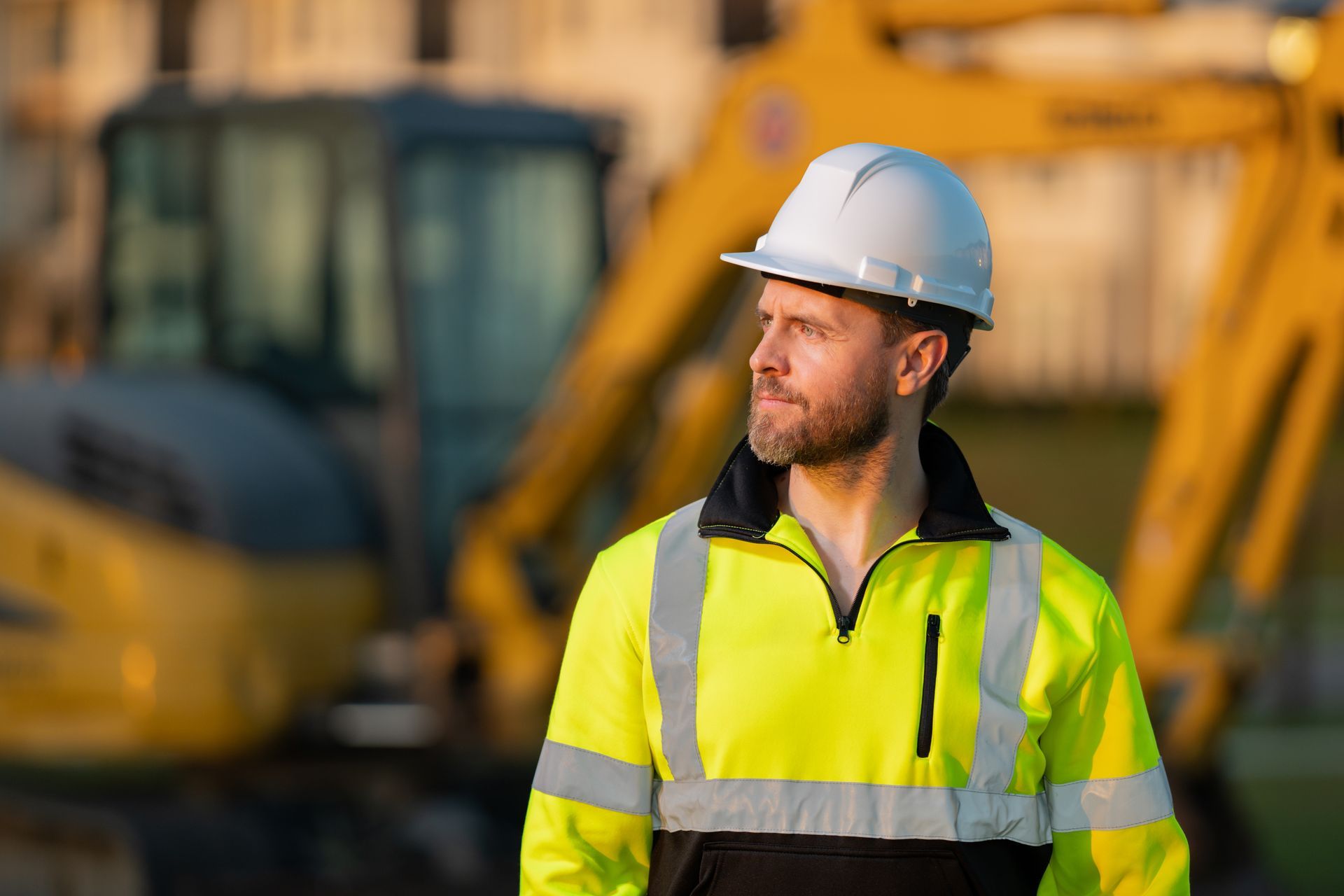 Construction worker in a hard hat and high-visibility jacket at a site with machinery behind.