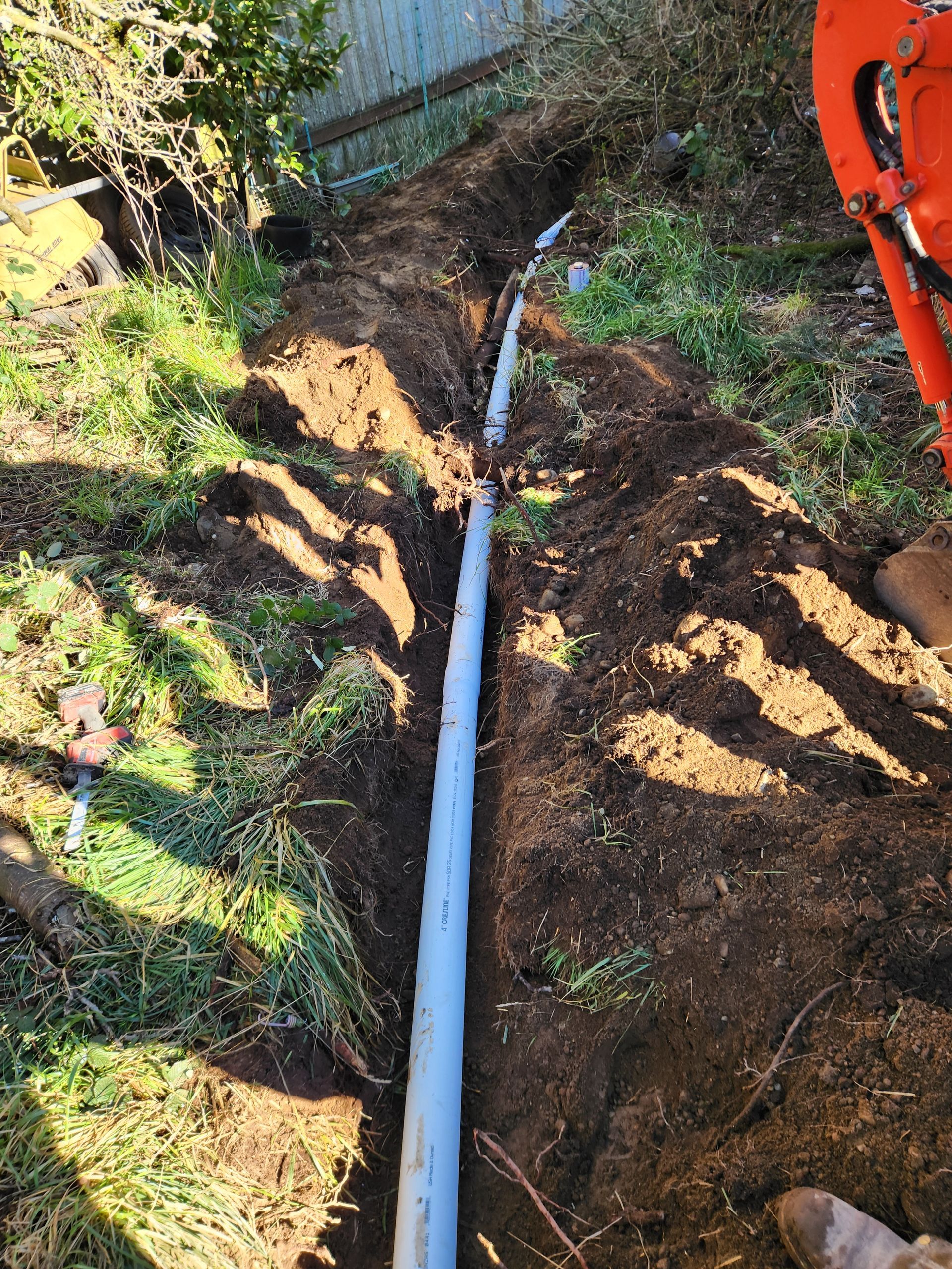 A grey pipe runs through a trench dug in a grassy yard, near an orange excavator.
