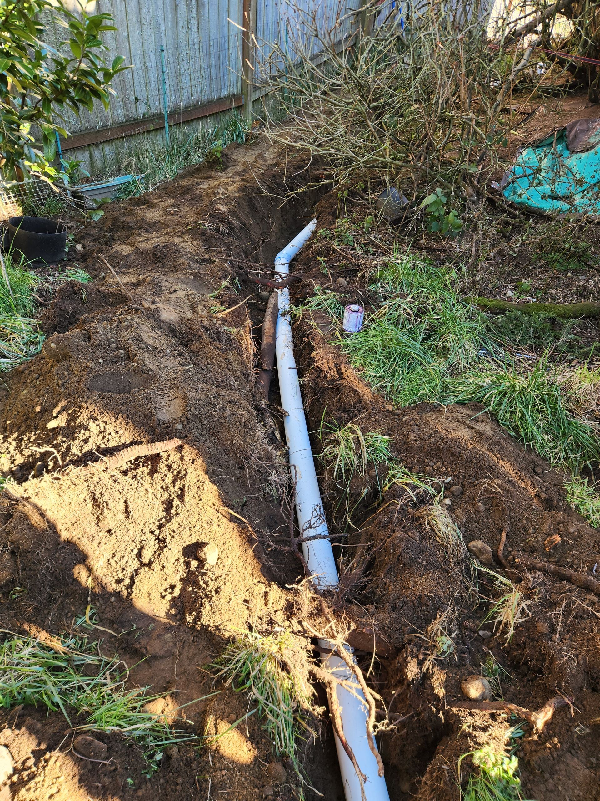 Grey drainage pipe in a trench, next to a brown dirt area, with foliage in the background.