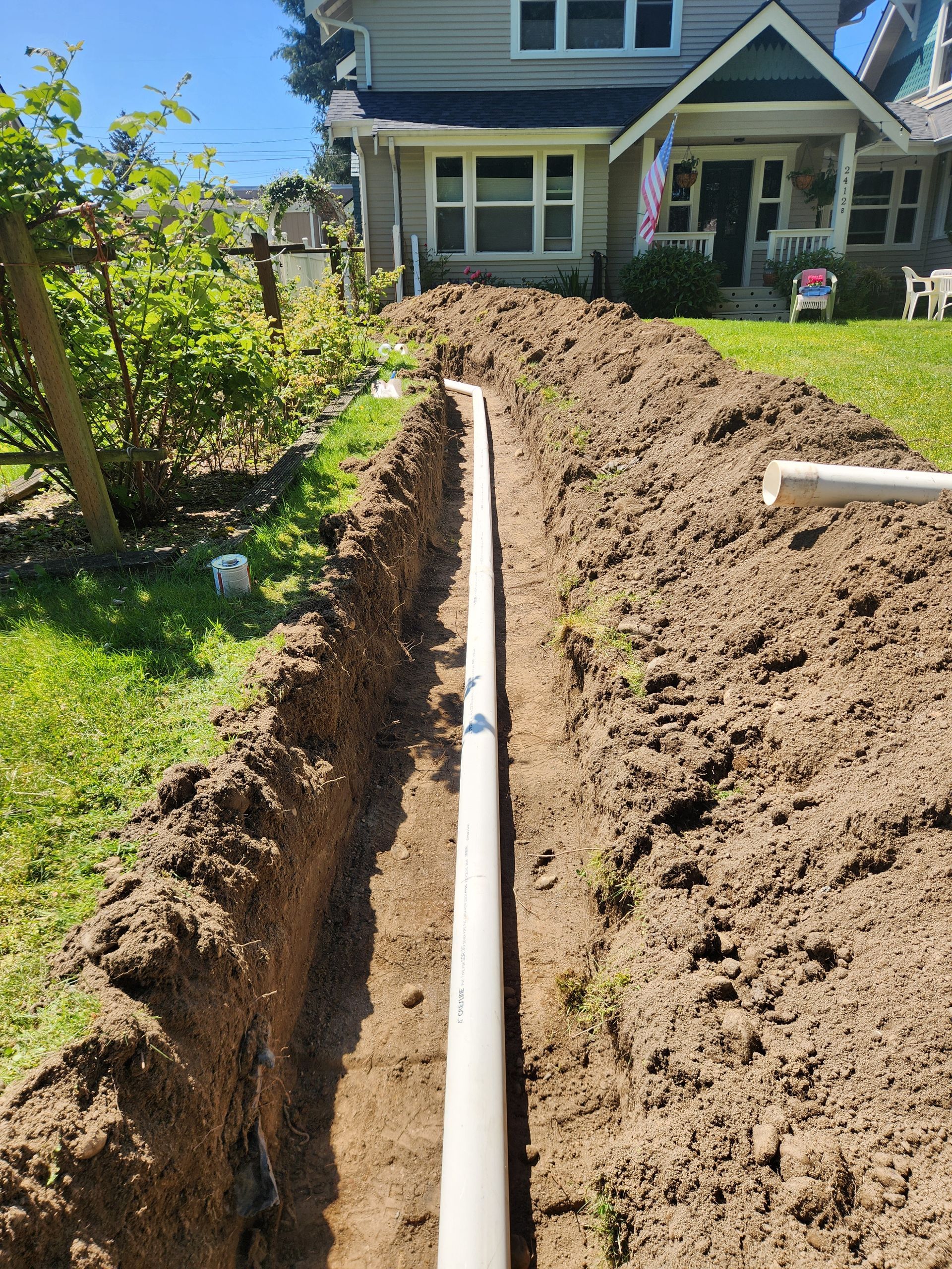 A long trench dug in a yard with a white pipe running through it in front of a house.