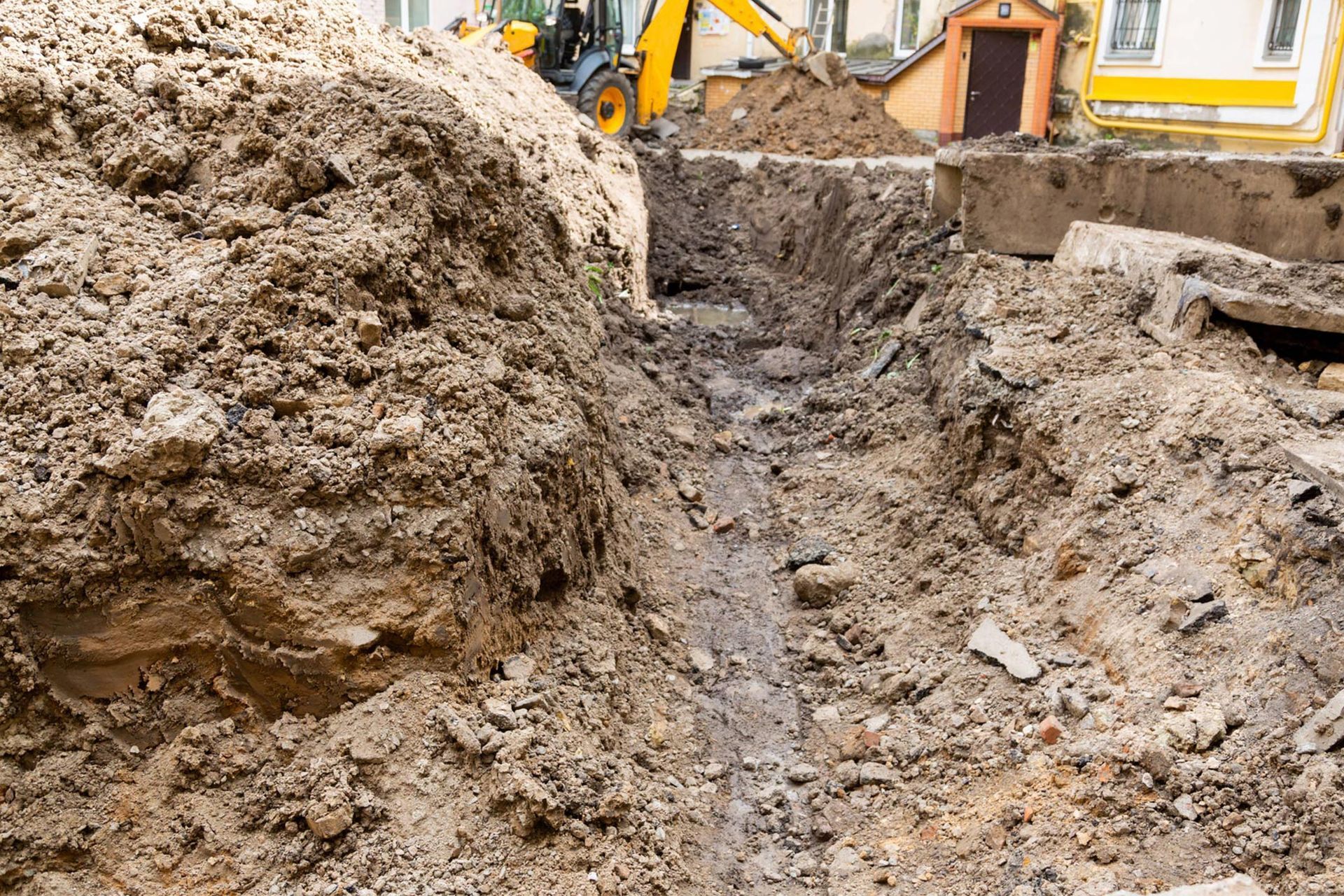 A large pile of dirt is being excavated by a bulldozer. Trench dug in dirt with an excavator in the background, near a building.