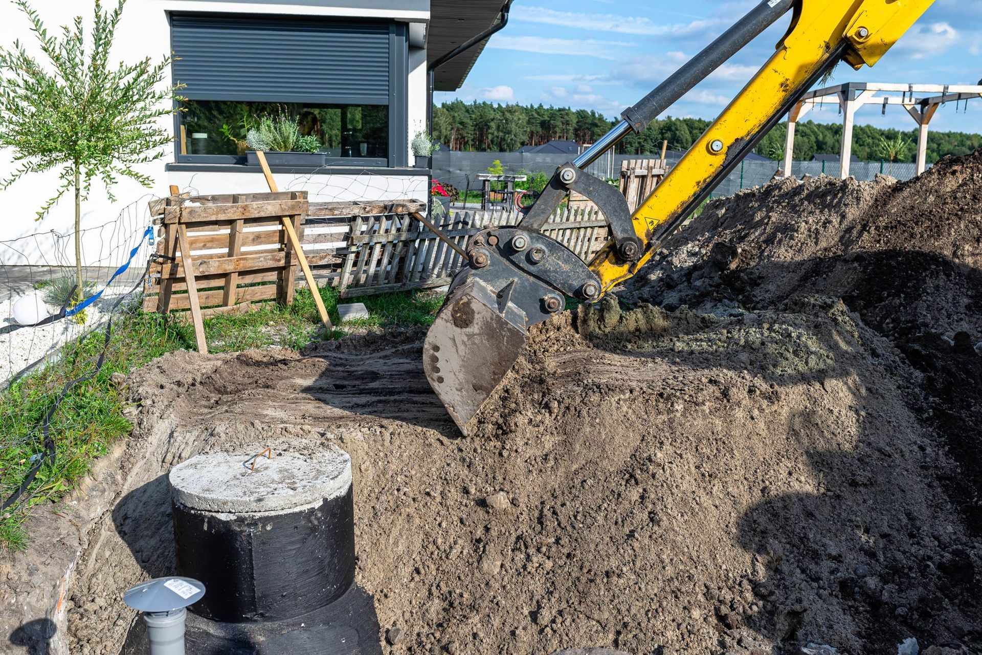 A yellow excavator is digging a hole in the ground in front of a house. A yellow excavator digs a hole in a yard, near a house and a buried septic tank.