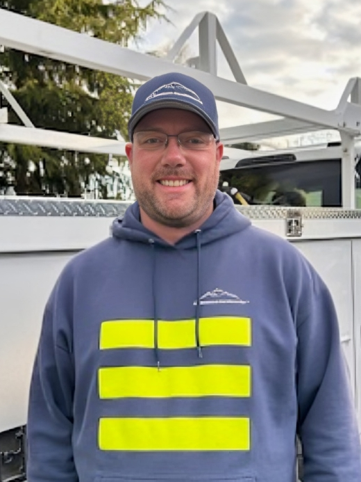 Man in blue hoodie and cap smiles in front of a white truck, logo on cap and hoodie.