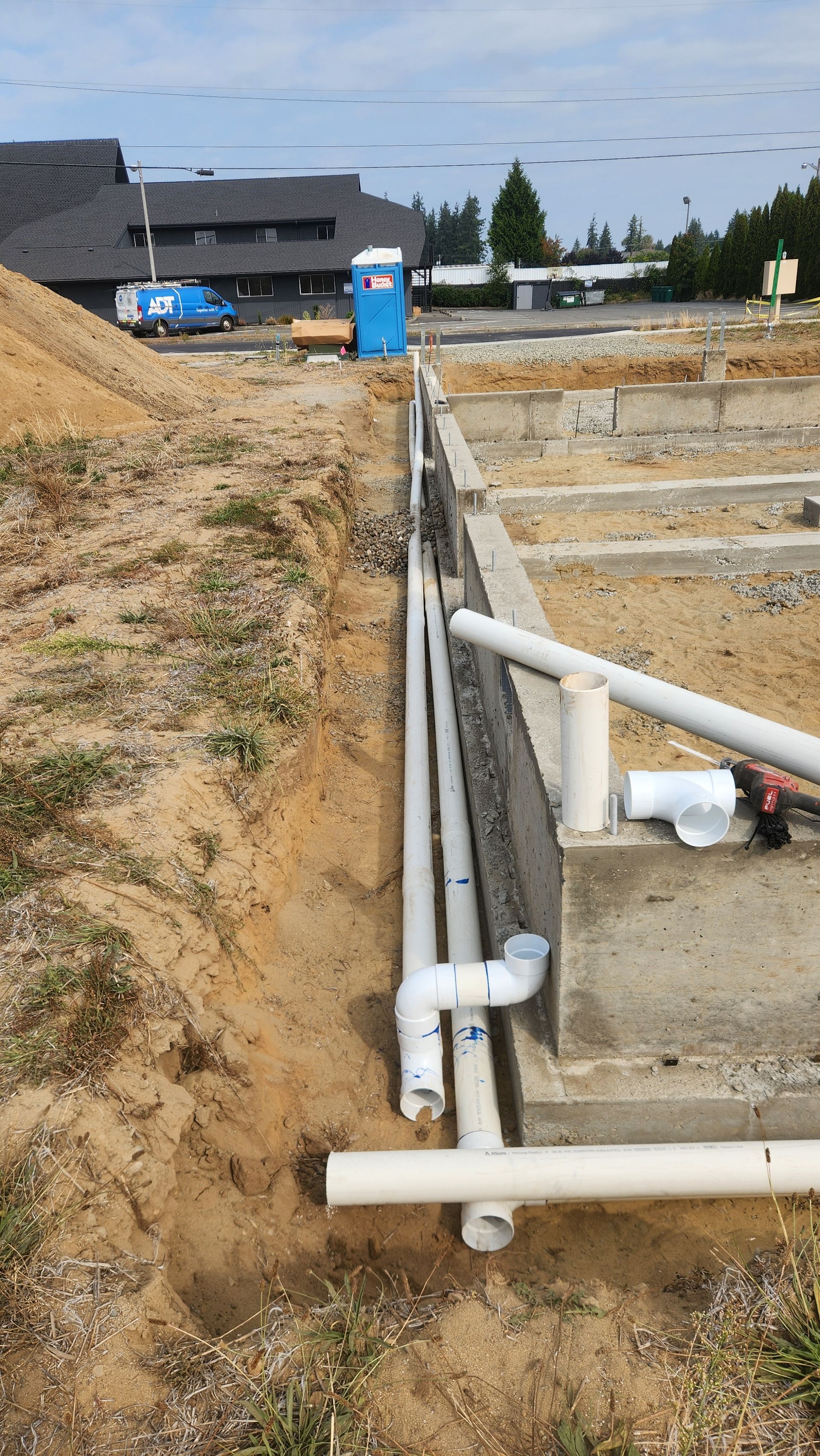 Construction site with trenches, concrete foundation, and PVC pipes. Blue porta-potty in background.