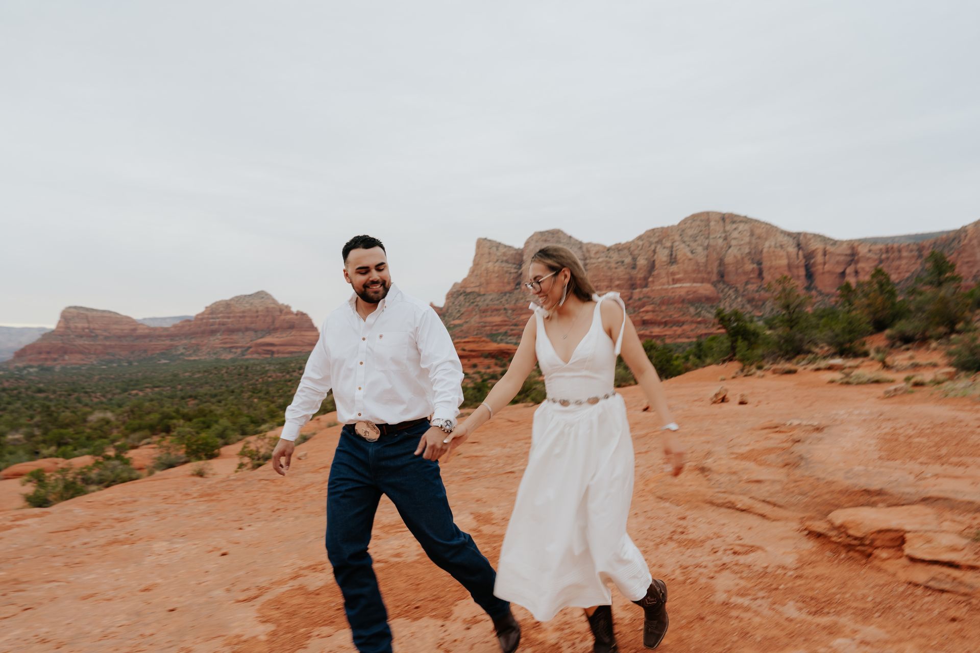 Couple holds hands, laughing, walking on red rock in Sedona, Arizona, with red rock formations in the background.