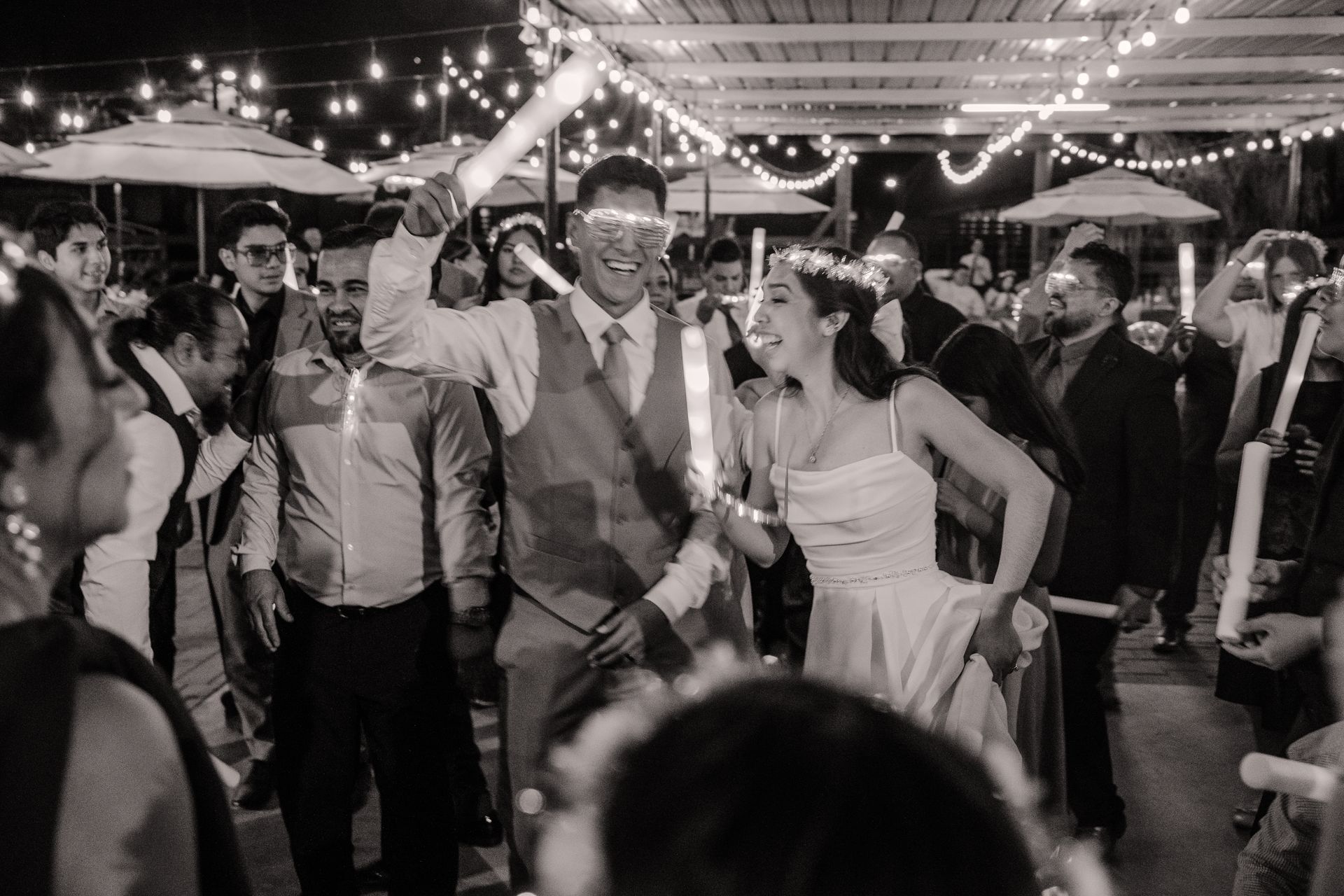 Newlyweds dance at reception, surrounded by guests holding glowing sticks. Outdoor setting with string lights.