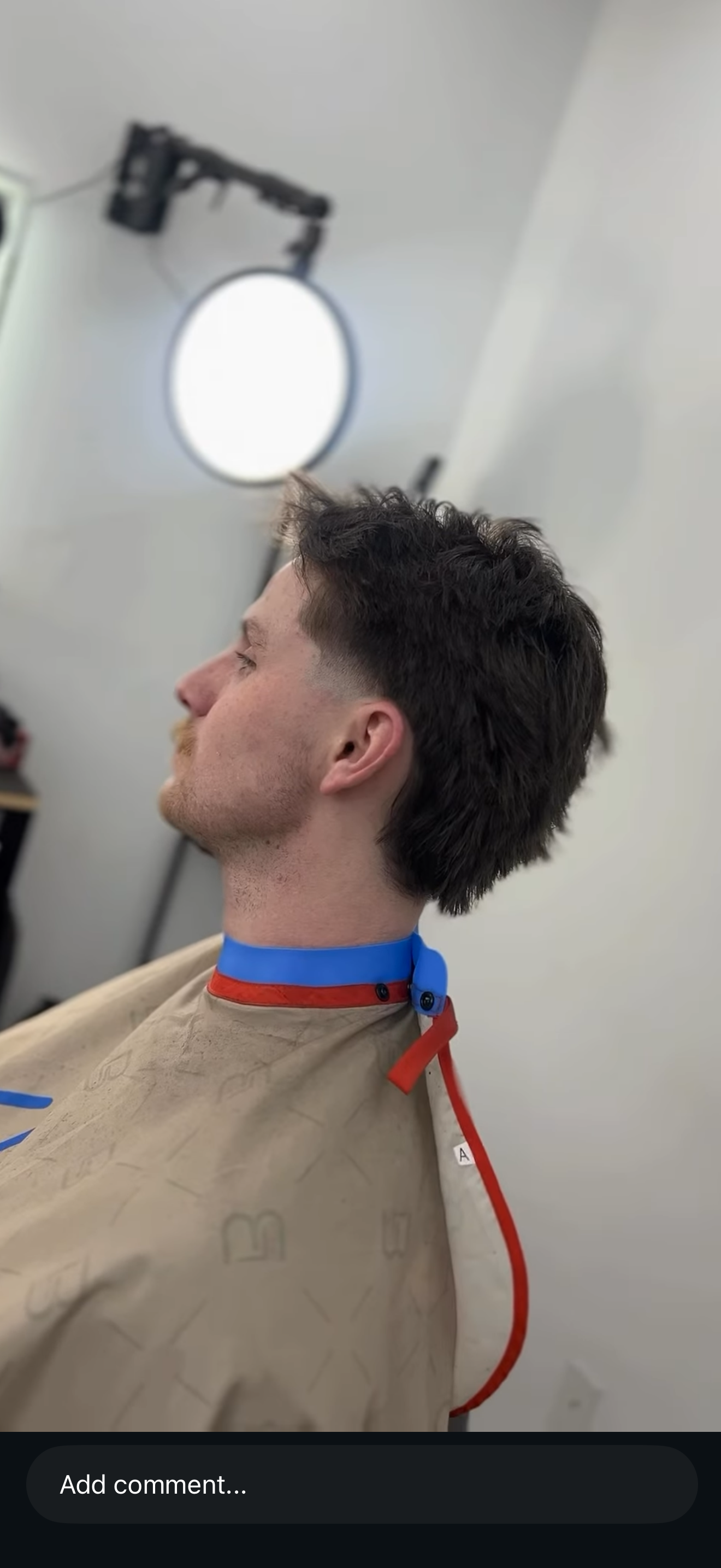 Side profile of a person wearing a beige shirt and blue-and-red neck strap, with a round wall light behind them.