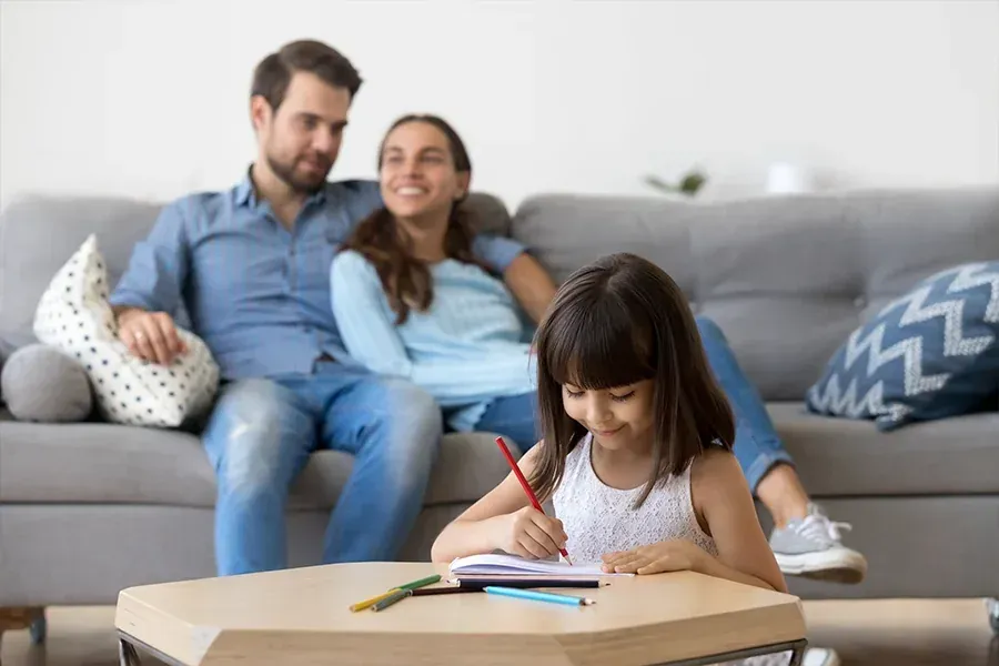 Child drawing at a table while two adults relax on a couch in the background.