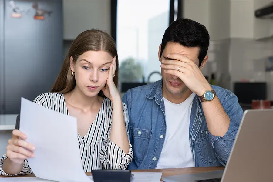 Two coworkers sit at a desk, looking stressed while reviewing paperwork beside a laptop.