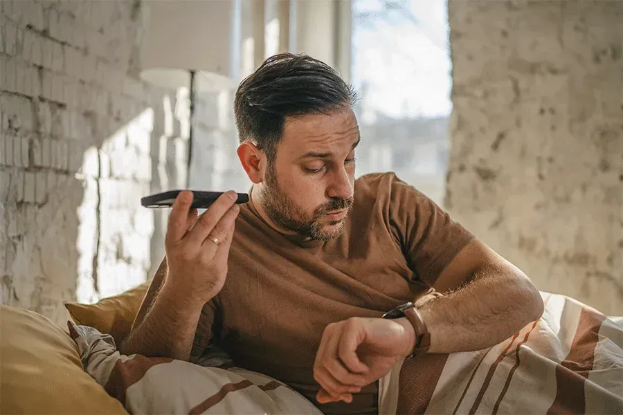 Man in brown sweater lying on a couch, looking at his smartwatch in a sunlit room