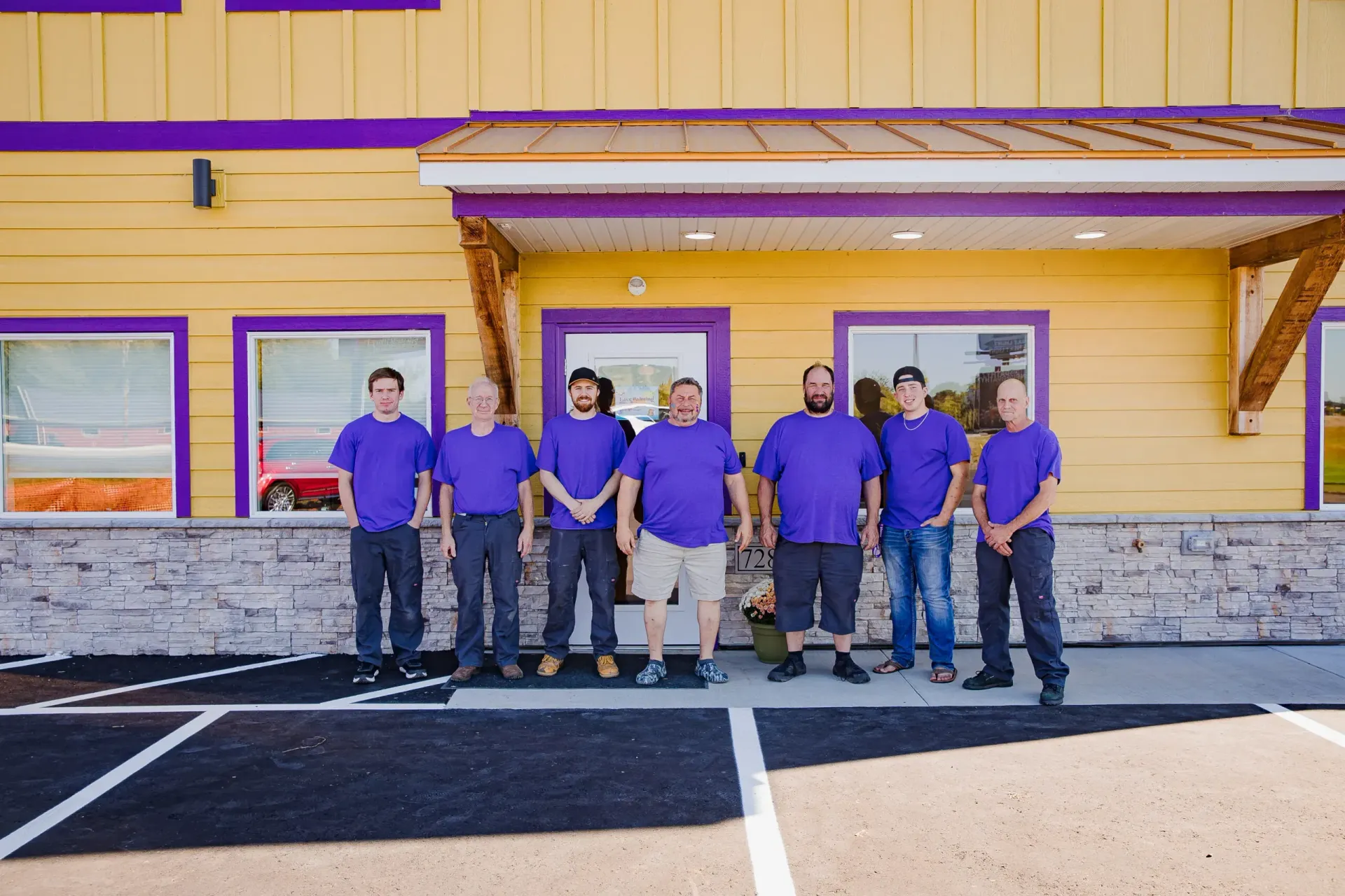 Six people in blue shirts standing outside a yellow building with stone trim and striped parking lines