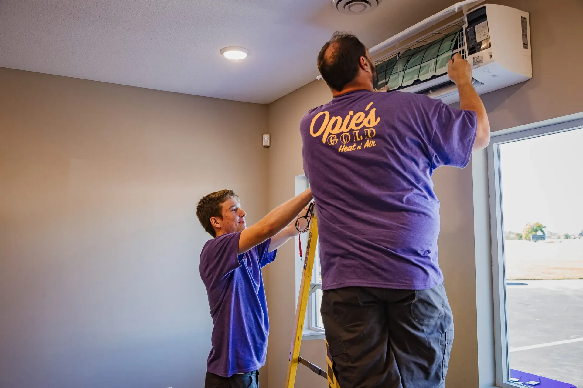 Two workers install a wall-mounted air conditioner in a room with a window and beige walls.