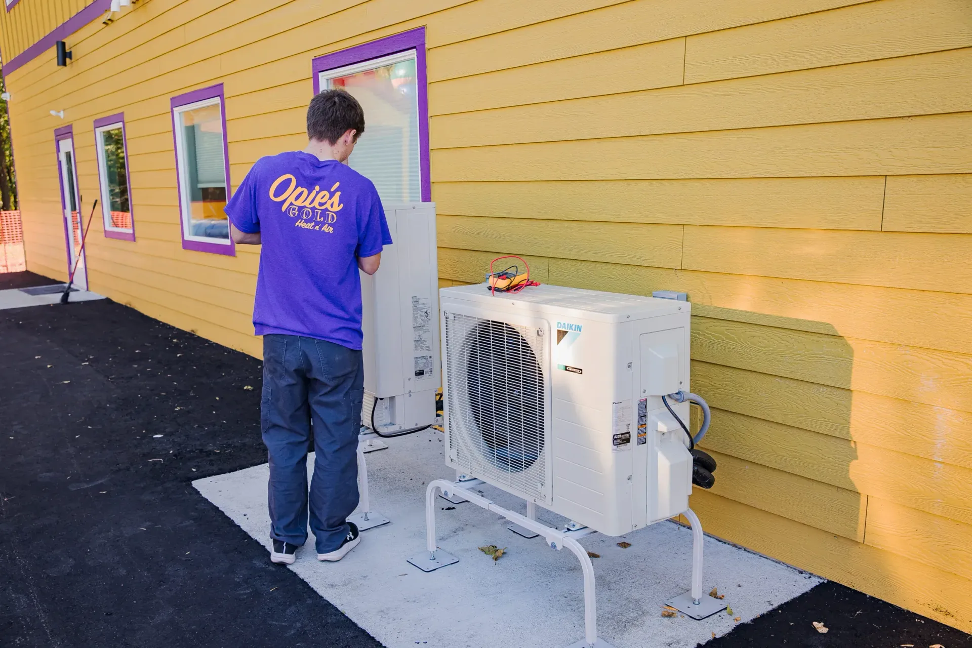 Technician standing by a wall-mounted outdoor HVAC unit beside a yellow building.