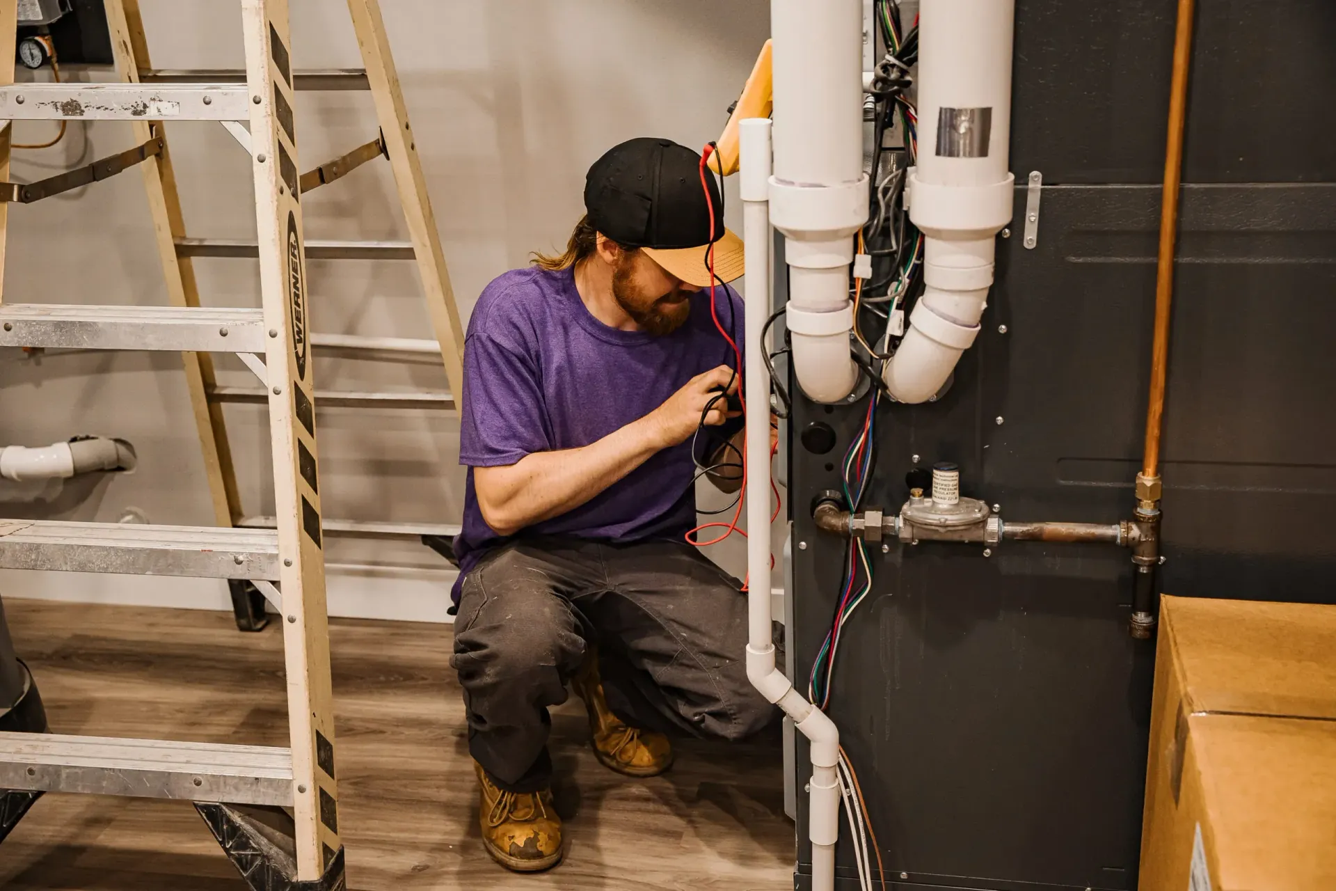 Worker in a purple shirt inspecting pipes in a utility room beside a ladder