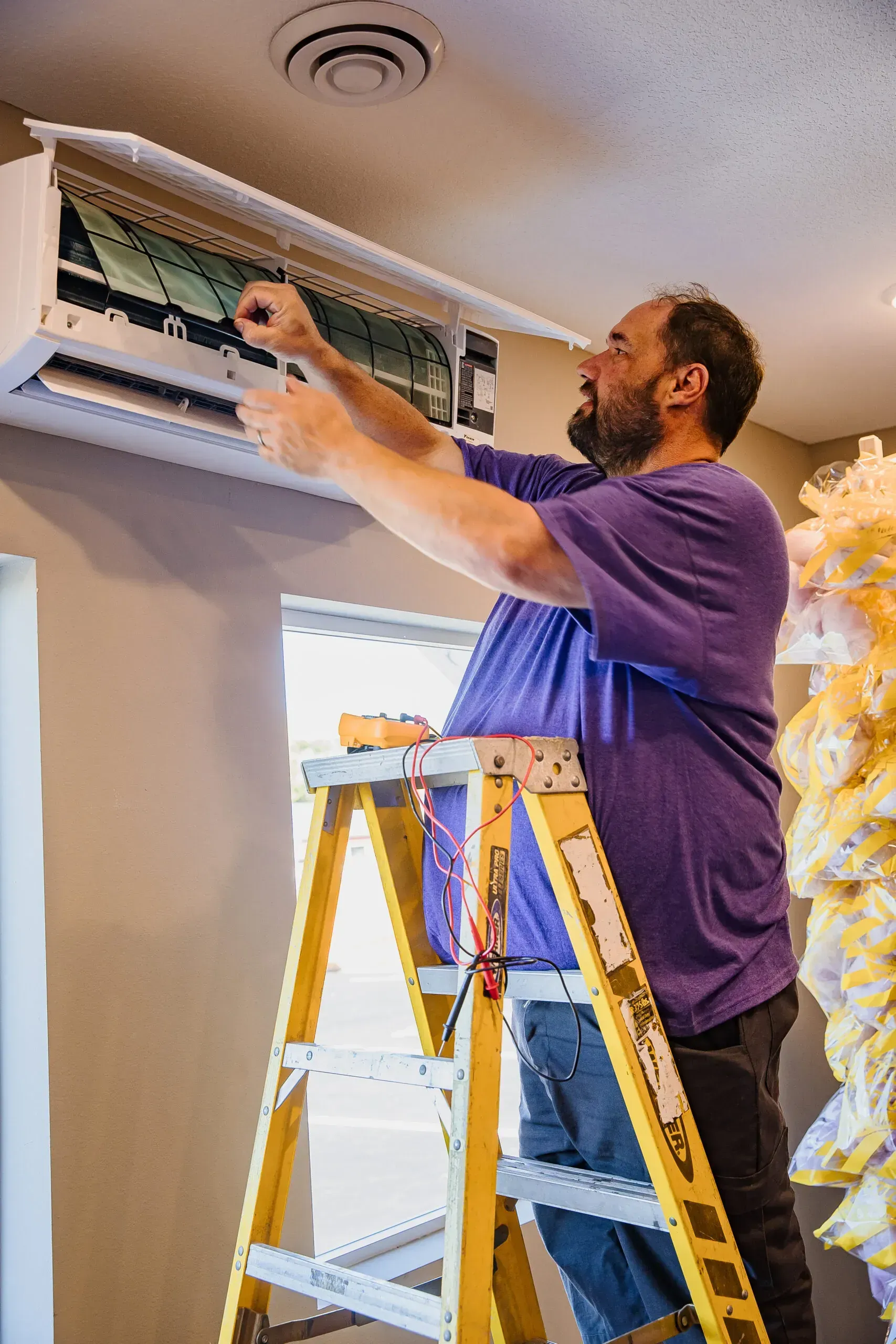Man in a purple shirt working on an air vent from a step ladder indoors