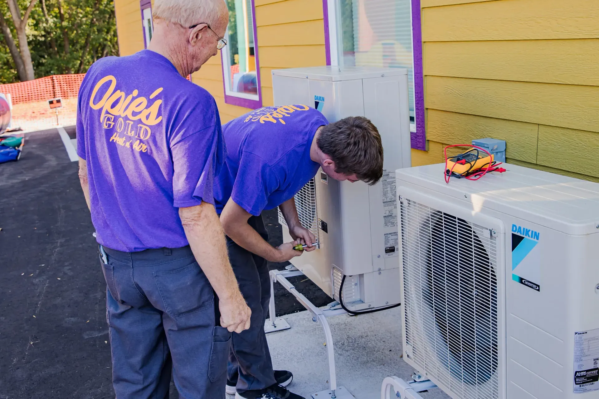 Two men in purple shirts work on outdoor air-conditioning units beside a yellow building.