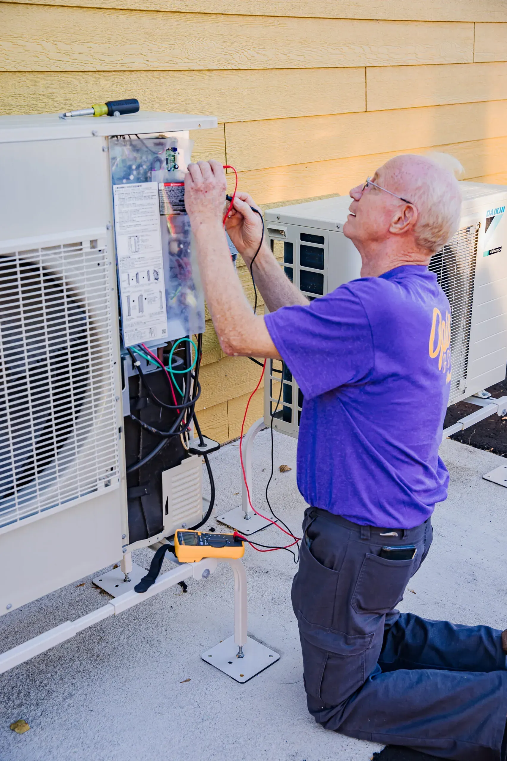 Technician kneeling beside HVAC units, inspecting wiring and notes on the wall outdoors