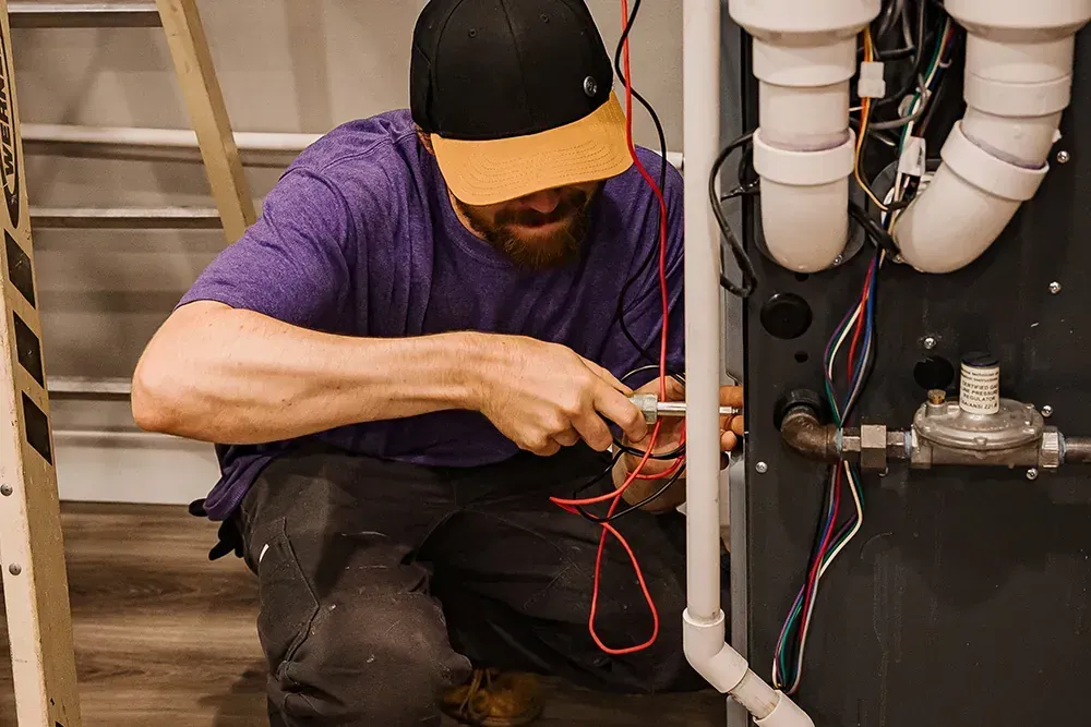 Worker in purple shirt and cap repairing plumbing under a sink with exposed pipes and tools