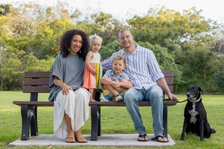 Family sitting on a park bench with a black dog nearby, trees and grass in the background.