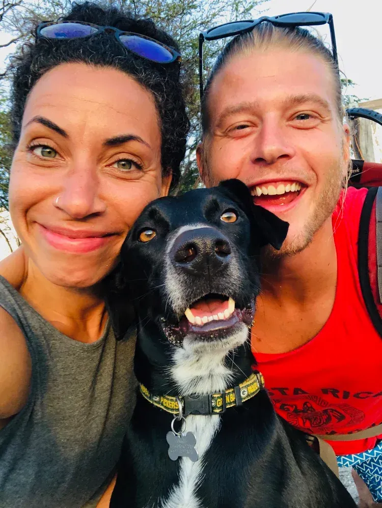 Two smiling people with a black dog in a close-up selfie outdoors