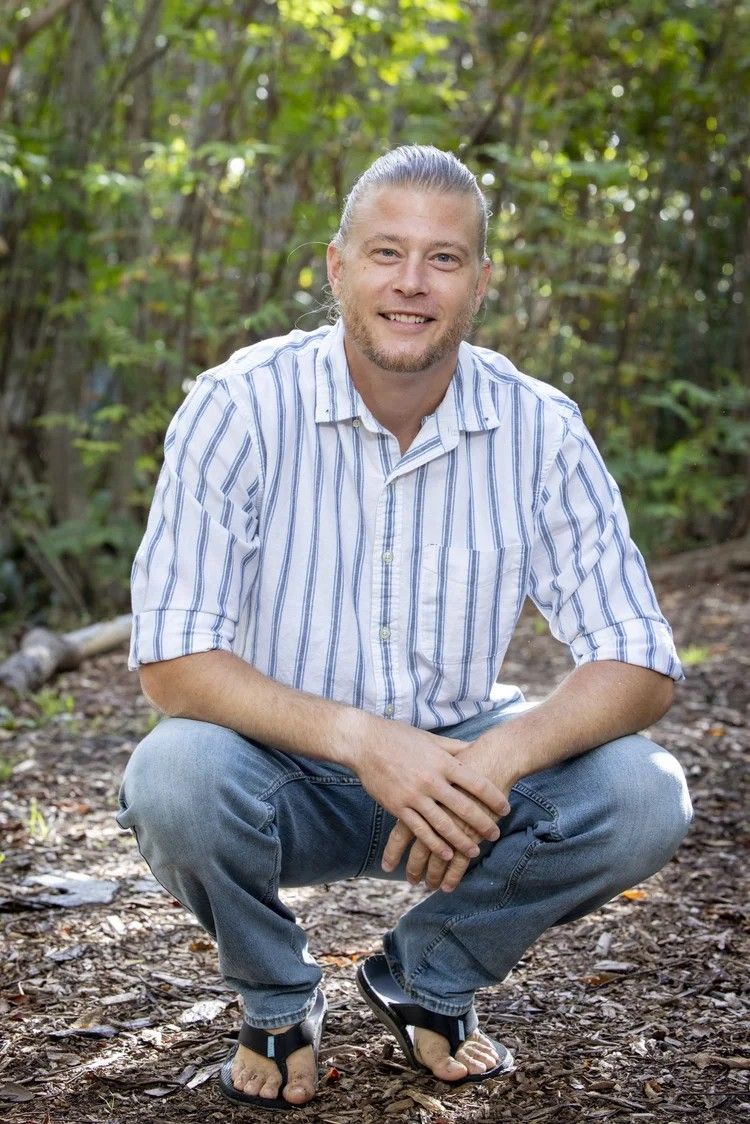 Man crouching outdoors on a forest trail, wearing a striped shirt and jeans, smiling at the camera