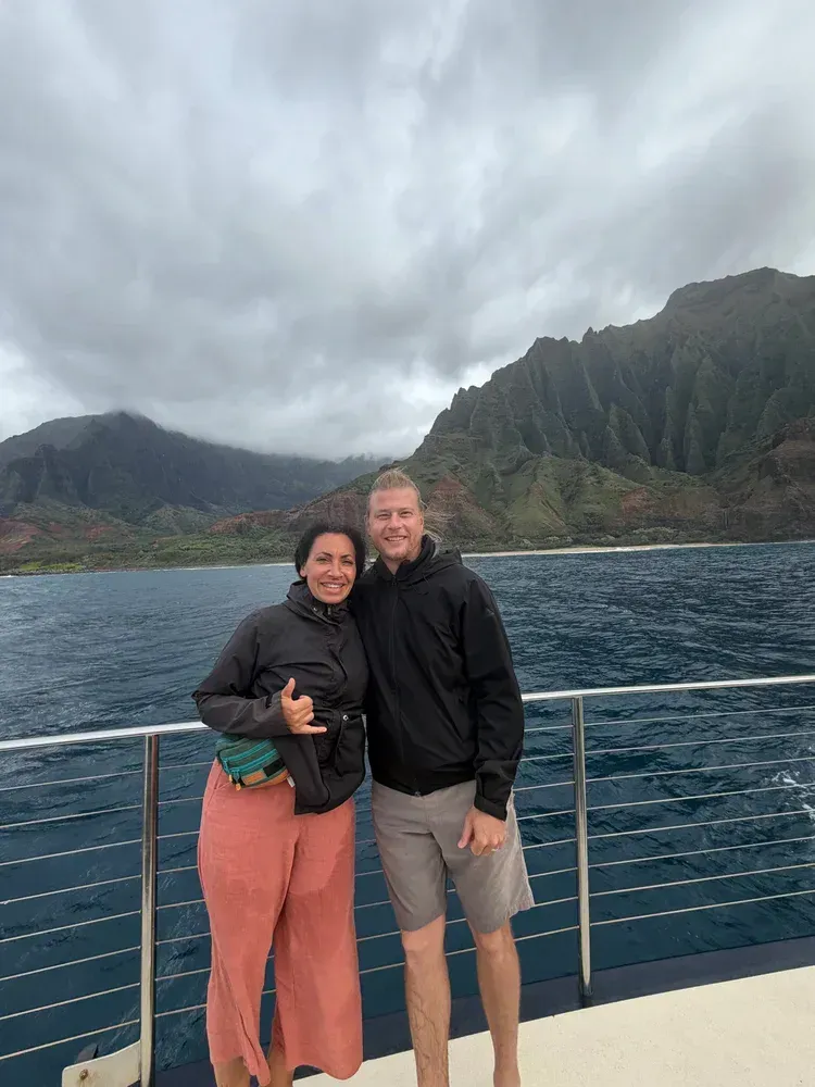 Two people standing on a boat deck with ocean and steep green mountains under cloudy skies.