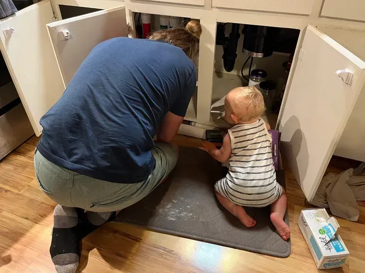 Adult and baby kneeling at an open cabinet, reaching toward items under the sink in a kitchen.