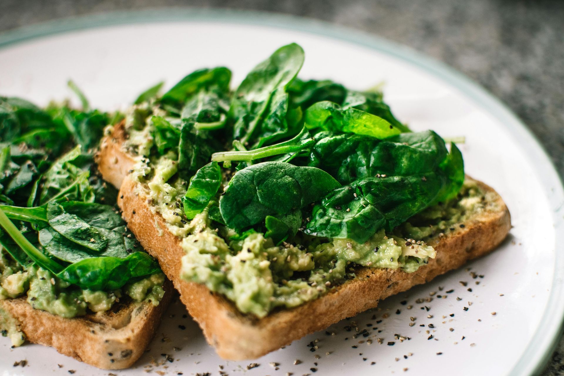 Two open-faced spinach toast slices on a white plate, sprinkled with black pepper