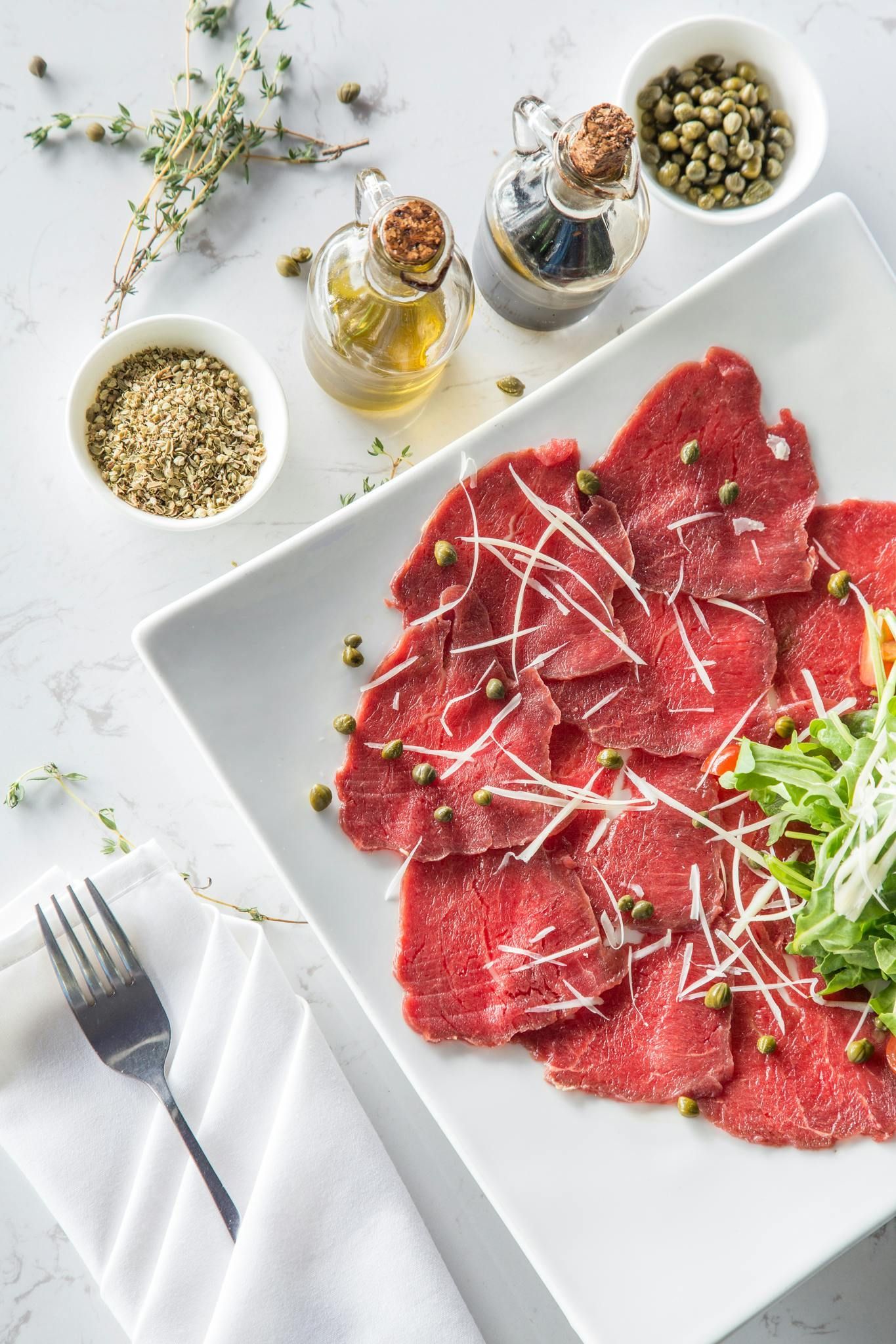 Thinly sliced raw beef carpaccio on a white plate with herbs, olive oil, and seasoning bowls