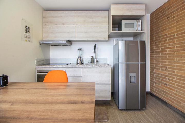Small modern kitchen with wood cabinets, stainless fridge, orange chair, and brick accent wall