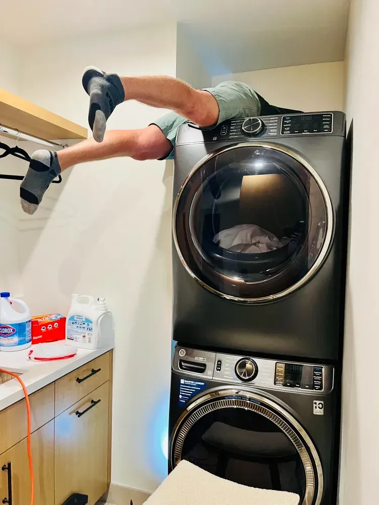 Person lying across stacked washer and dryer in a laundry room, legs hanging off the side.