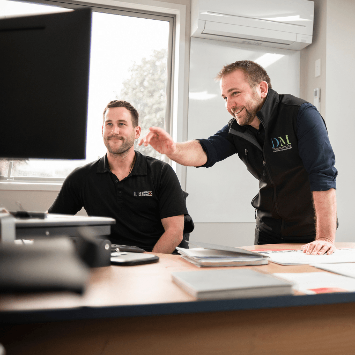 Two coworkers smiling and discussing work in a bright office, one pointing at a computer screen