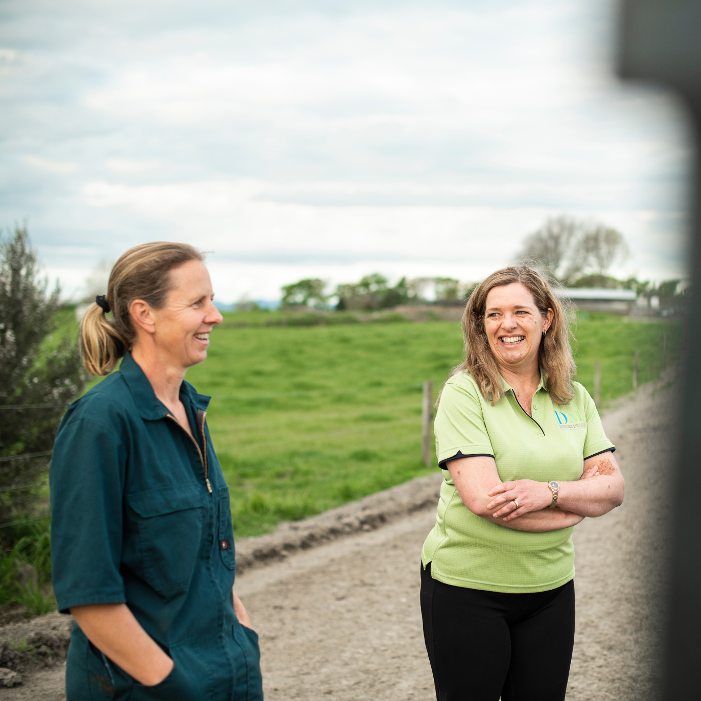 Two women standing and smiling on a dirt path beside a green field under a cloudy sky