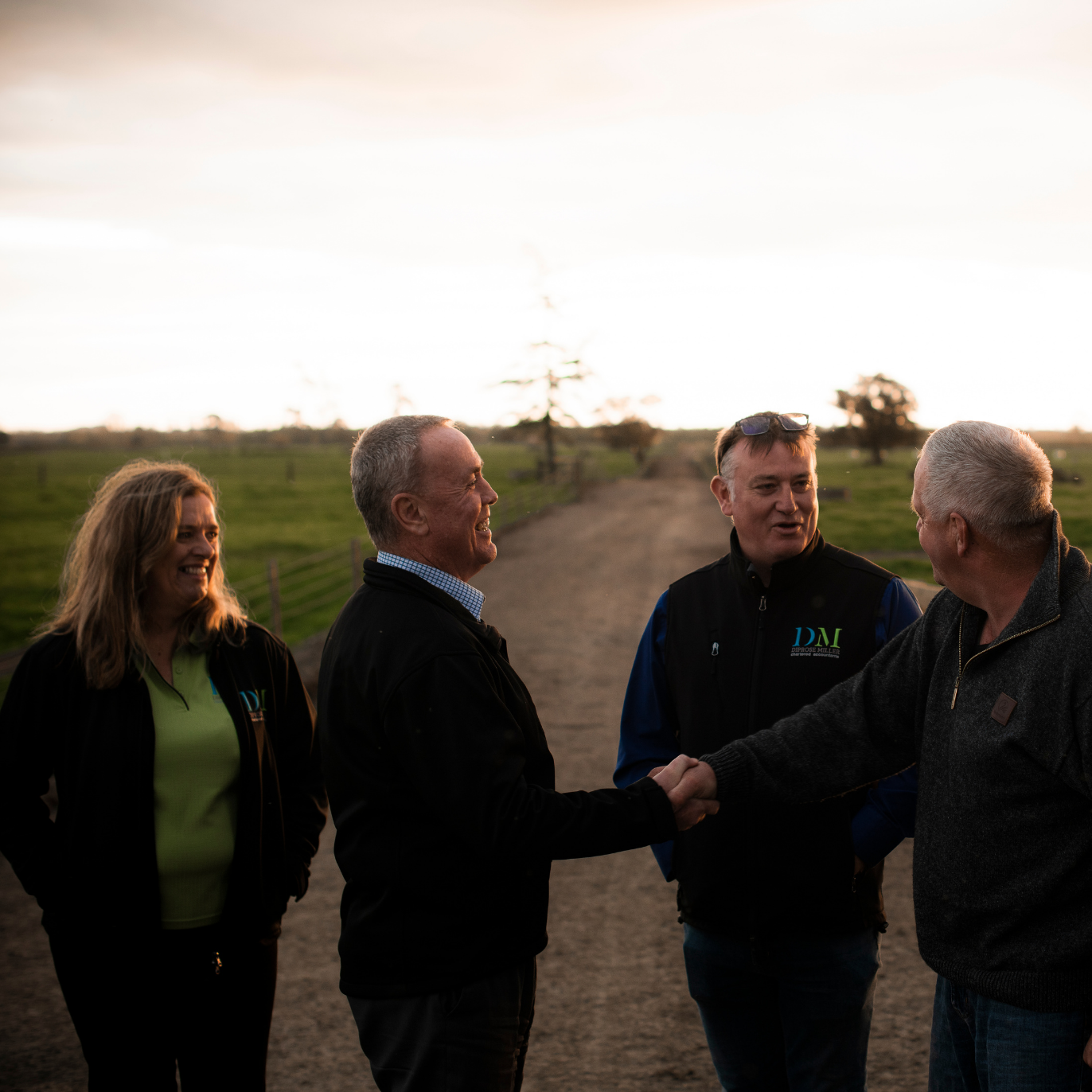 Four people shake hands on a rural road at sunset, with fields and a cloudy sky behind them.