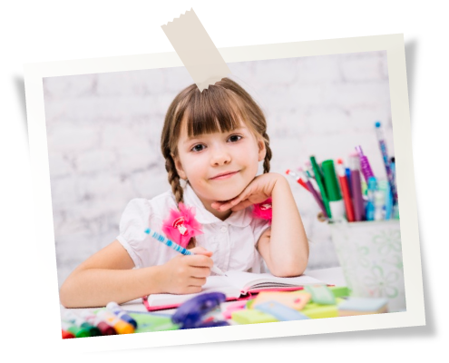 A young girl with pigtails smiles, leaning on her hand while writing at a desk with colorful art supplies.