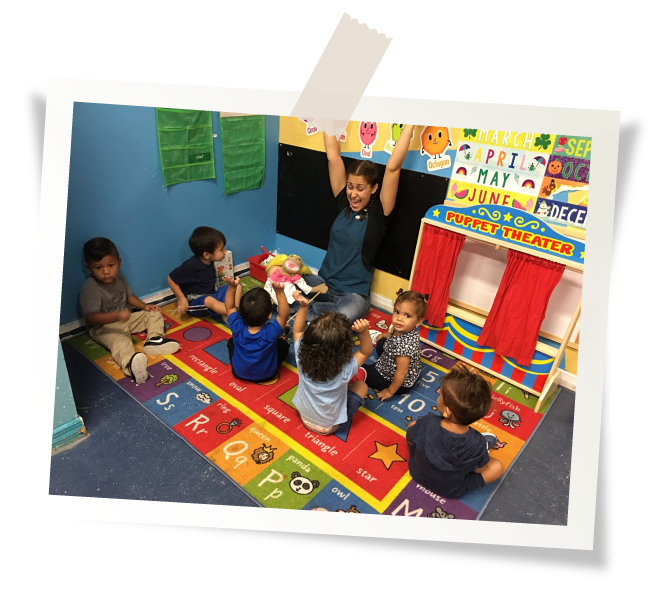 Children and teacher in a classroom, gathered on a colorful rug. The teacher raises her arms, and a puppet theater is visible.