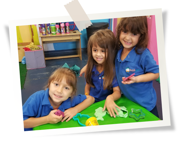 Three young girls in blue shirts smile while playing with colorful clay at a green table in a classroom.