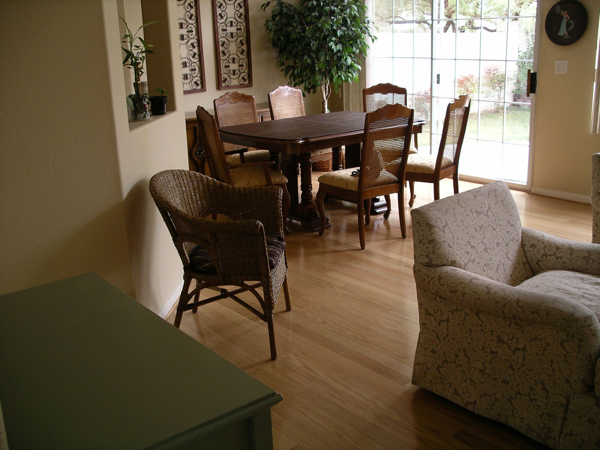 Sunlit dining room with wooden table and chairs, floral armchairs, and a green side table near a window