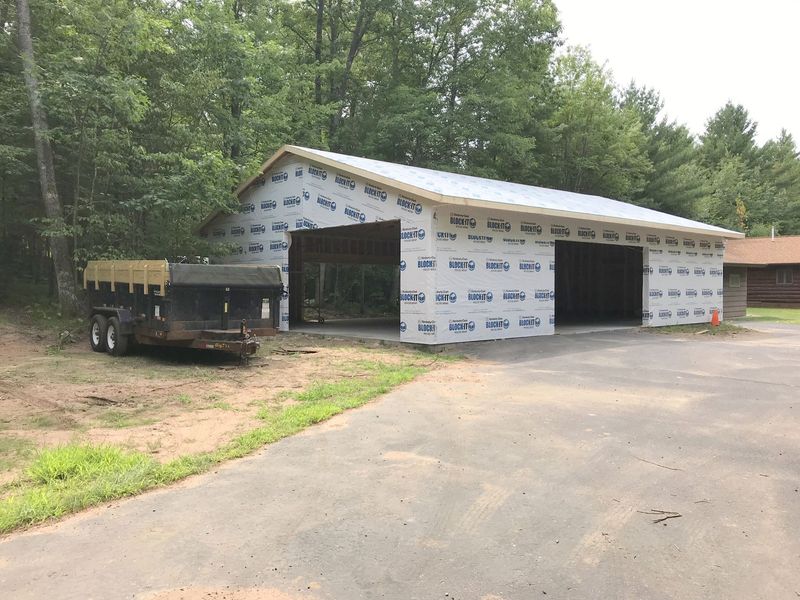 Wood-framed house under construction with wrapped exterior walls beside a wooded area