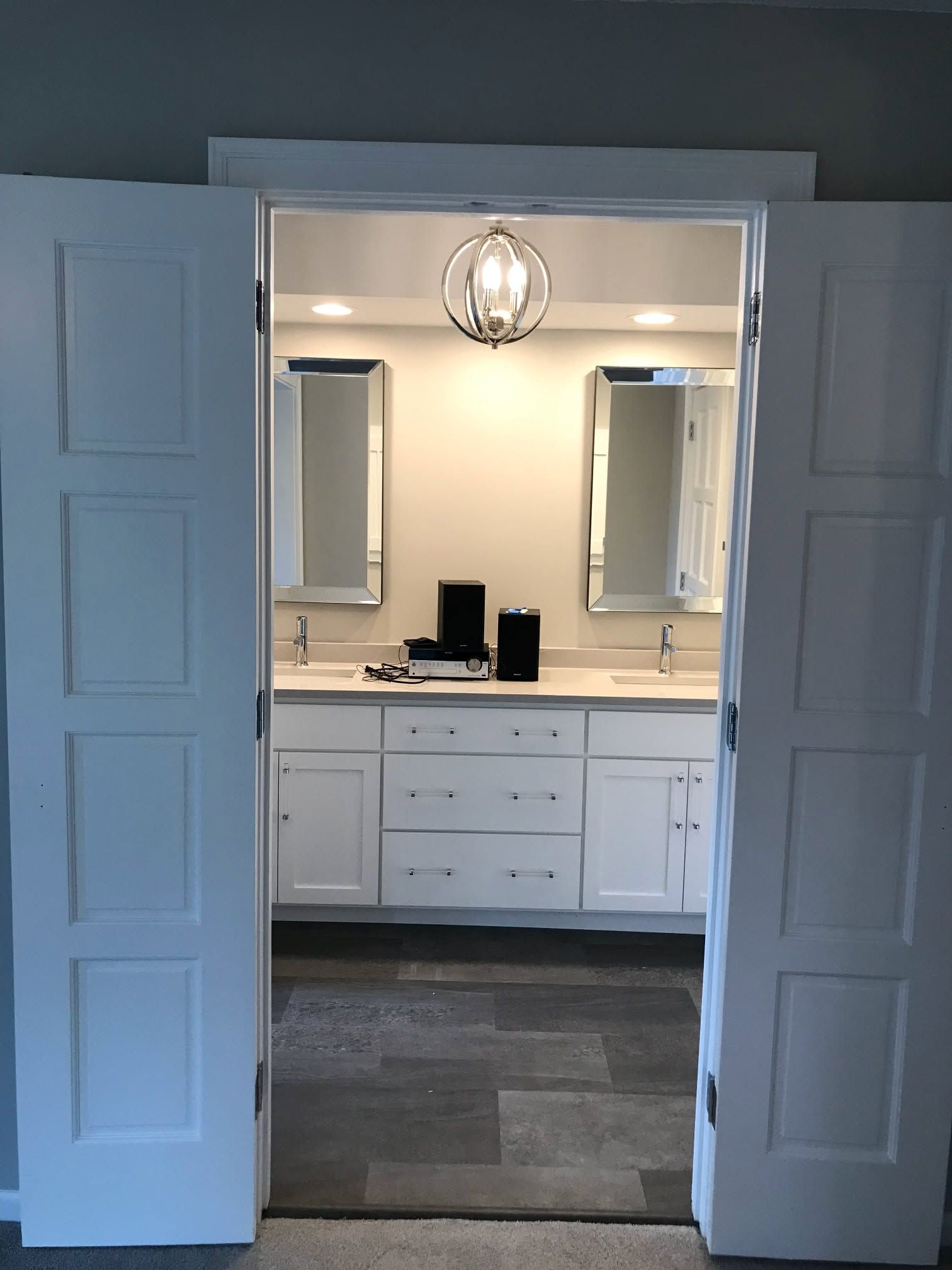 Open double doors to a bright bathroom with a double vanity, mirrors, and a hanging light fixture.