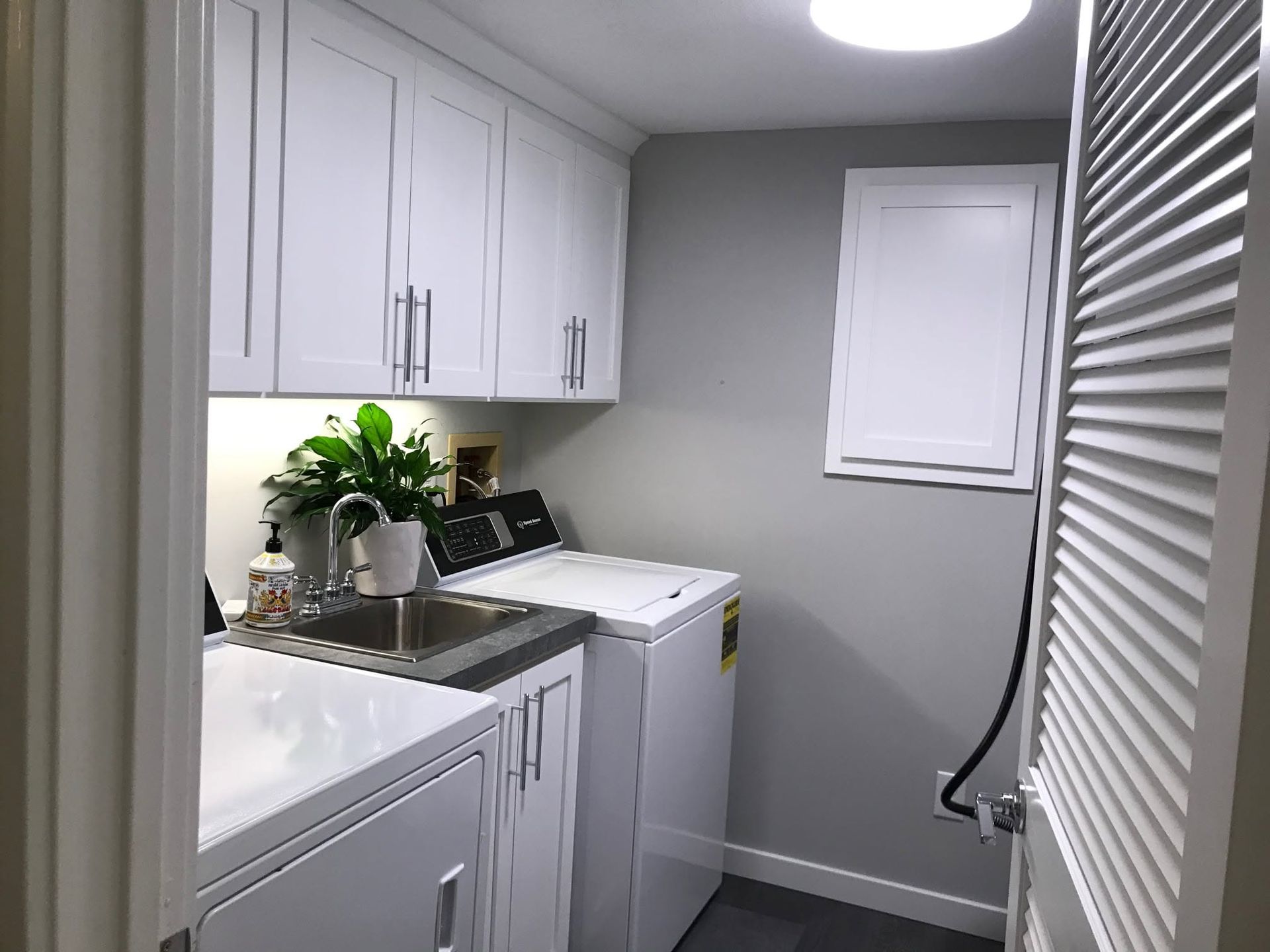 Small laundry room with white cabinets, sink, washing machine, and a window with blinds.