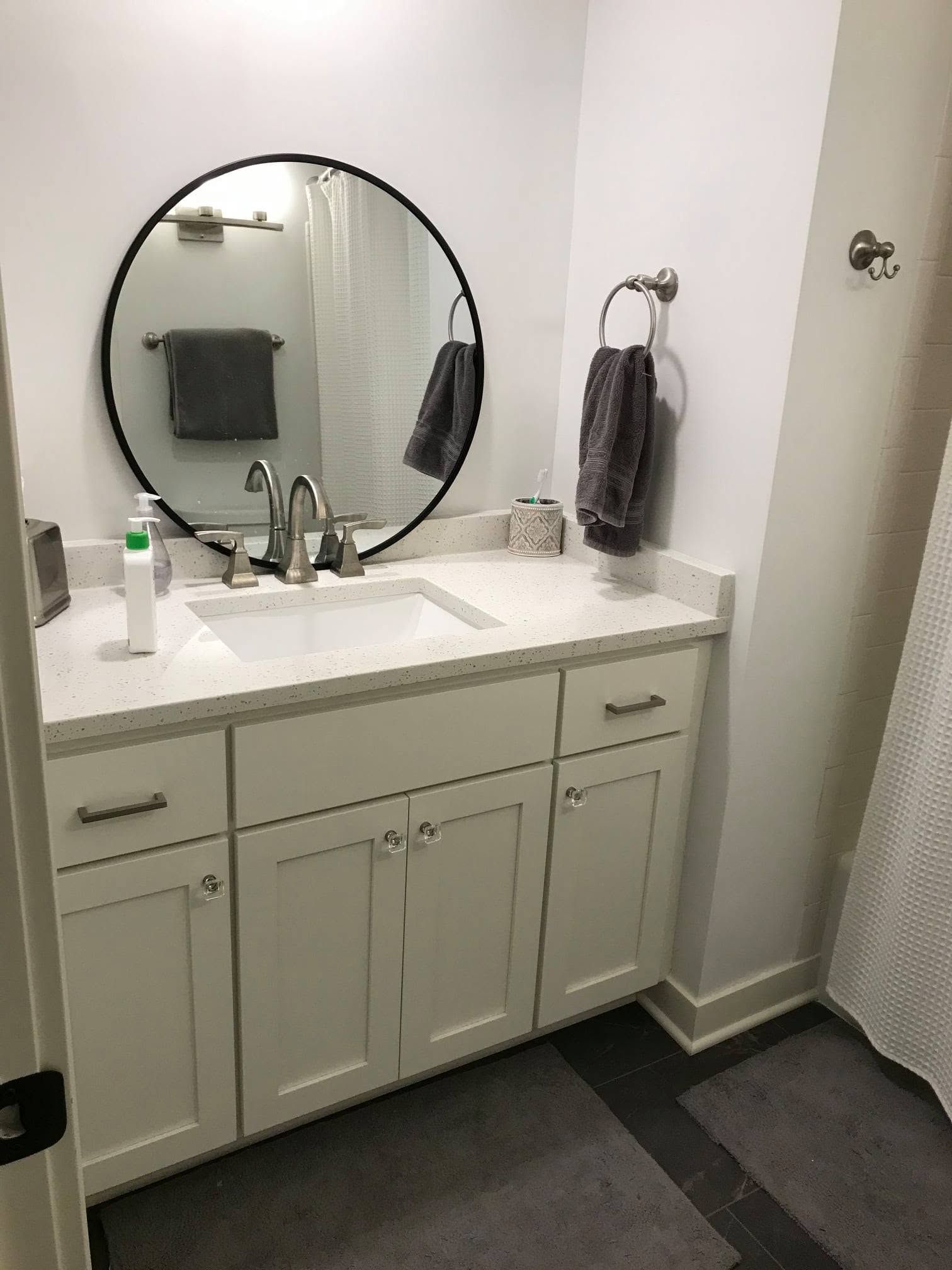 Small white bathroom vanity with round mirror, sink, towel ring, and dark floor mats