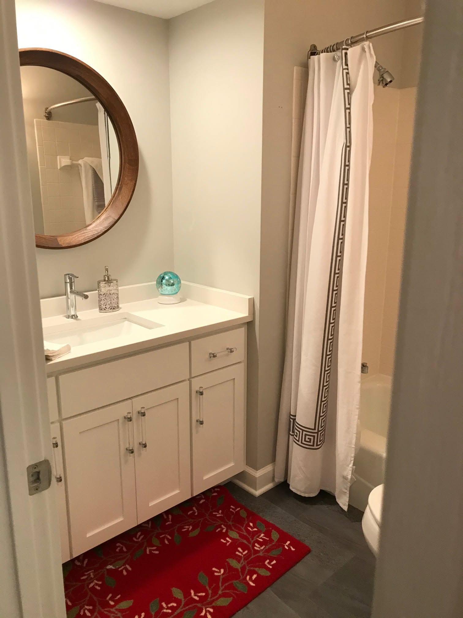 Small bathroom with white vanity, round mirror, shower curtain, and red patterned rug.