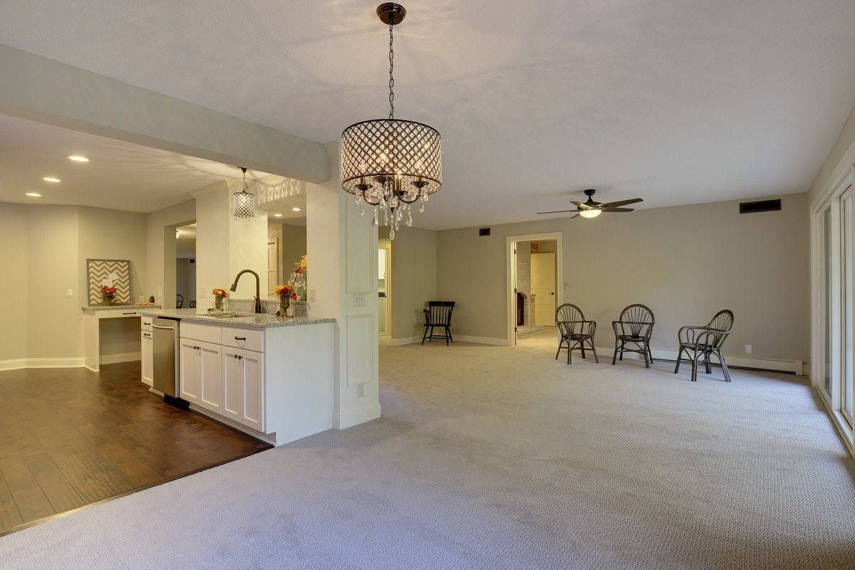 Spacious carpeted living area with white kitchen island, chandelier, and seating by a doorway.