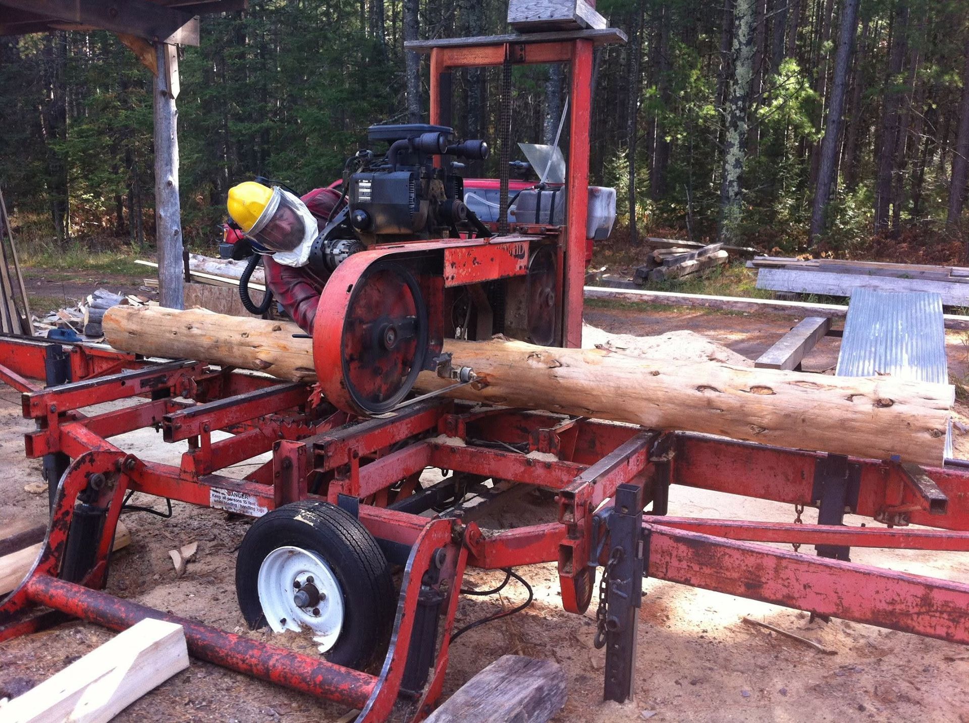 Portable sawmill cutting a log outdoors, with a worker in a yellow hard hat nearby.