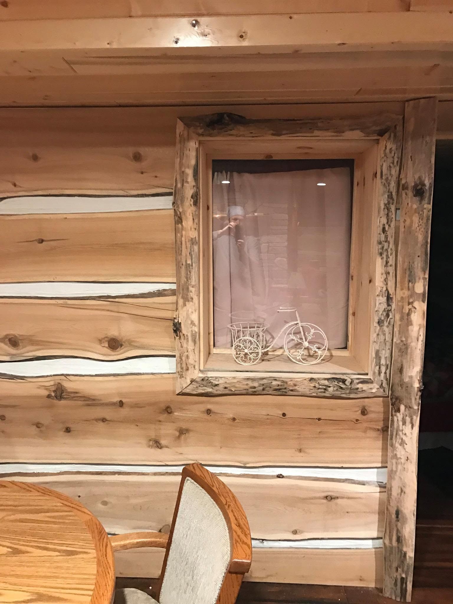 Rustic log cabin wall with a small curtained window and a wicker chair in the foreground