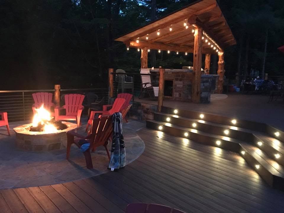 Backyard deck at dusk with a lit fire pit, red chairs, and a pergola bar with string lights and steps.