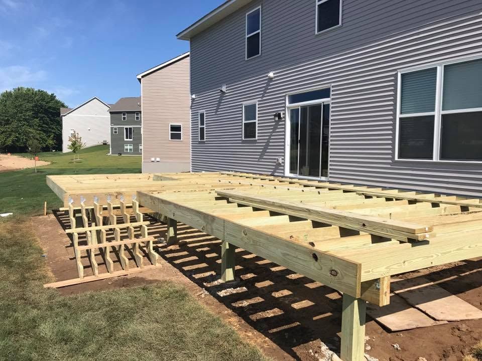 Backyard wooden deck frame under construction beside a gray house, with lumber supports and joists visible.